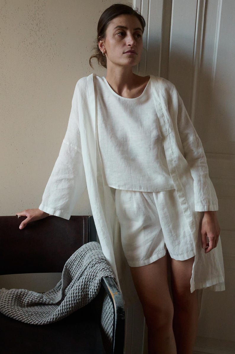 May include: A white linen loungewear set, including a long-sleeved top, shorts, and a long, open-front robe. The outfit is lightweight and appears comfortable. A gray textured blanket is draped over a dark brown chair.