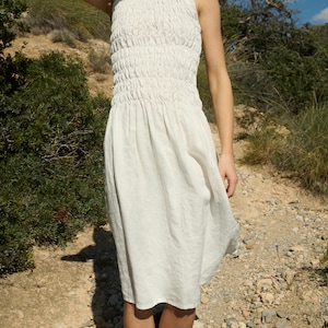 May include: A white sleeveless dress with a smocked bodice and a flowing skirt. The dress is worn by a person standing on a dirt path.