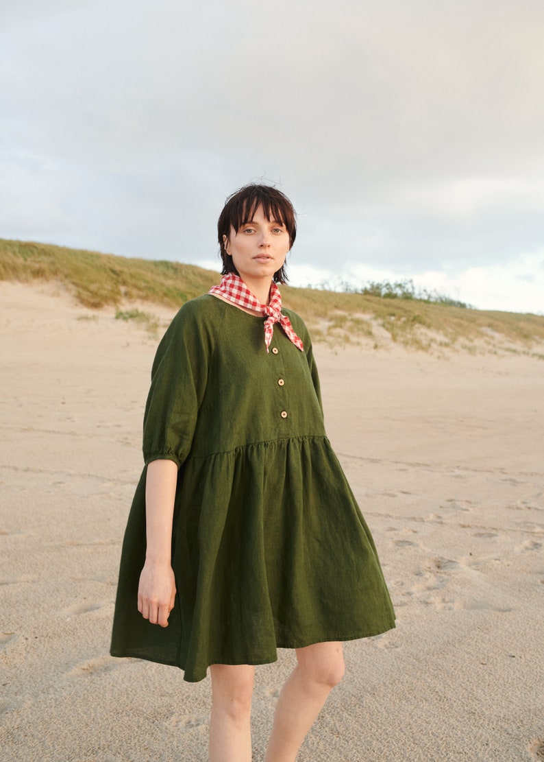 May include: A woman wearing a green linen dress with a red and white gingham scarf on a sandy beach.