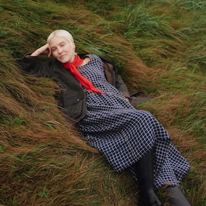 May include: A navy blue and white checked long dress worn with a dark olive green waxed jacket, a bright red scarf, and black rubber boots. The garment has a simple round neckline and a loose, flowing silhouette with long sleeves.