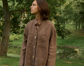May include: A brown linen jacket with a mandarin collar and wooden buttons. The jacket has two front pockets and is worn with black pants. The person is standing in a park with green grass and trees in the background.