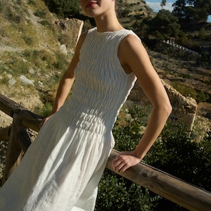May include: A woman wearing a white linen dress with a smocked bodice stands on a wooden railing overlooking a hillside.