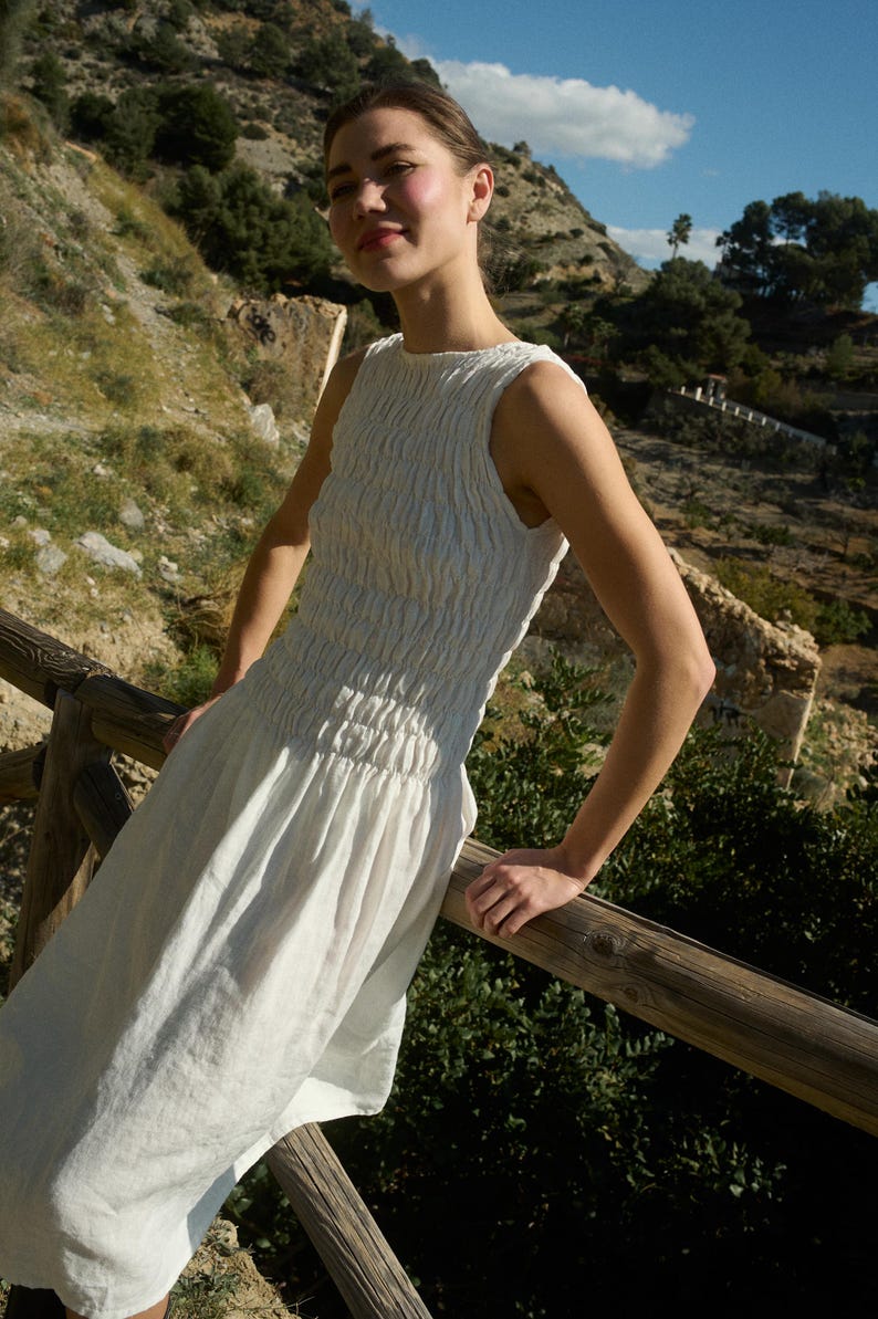 May include: A woman wearing a white linen dress with a smocked bodice stands on a wooden railing overlooking a hillside.
