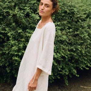 May include: A woman wearing a white linen dress with a square neckline and three-quarter sleeves. The dress has a ruffled hem and is made of a lightweight fabric. The background is a lush green hedge.