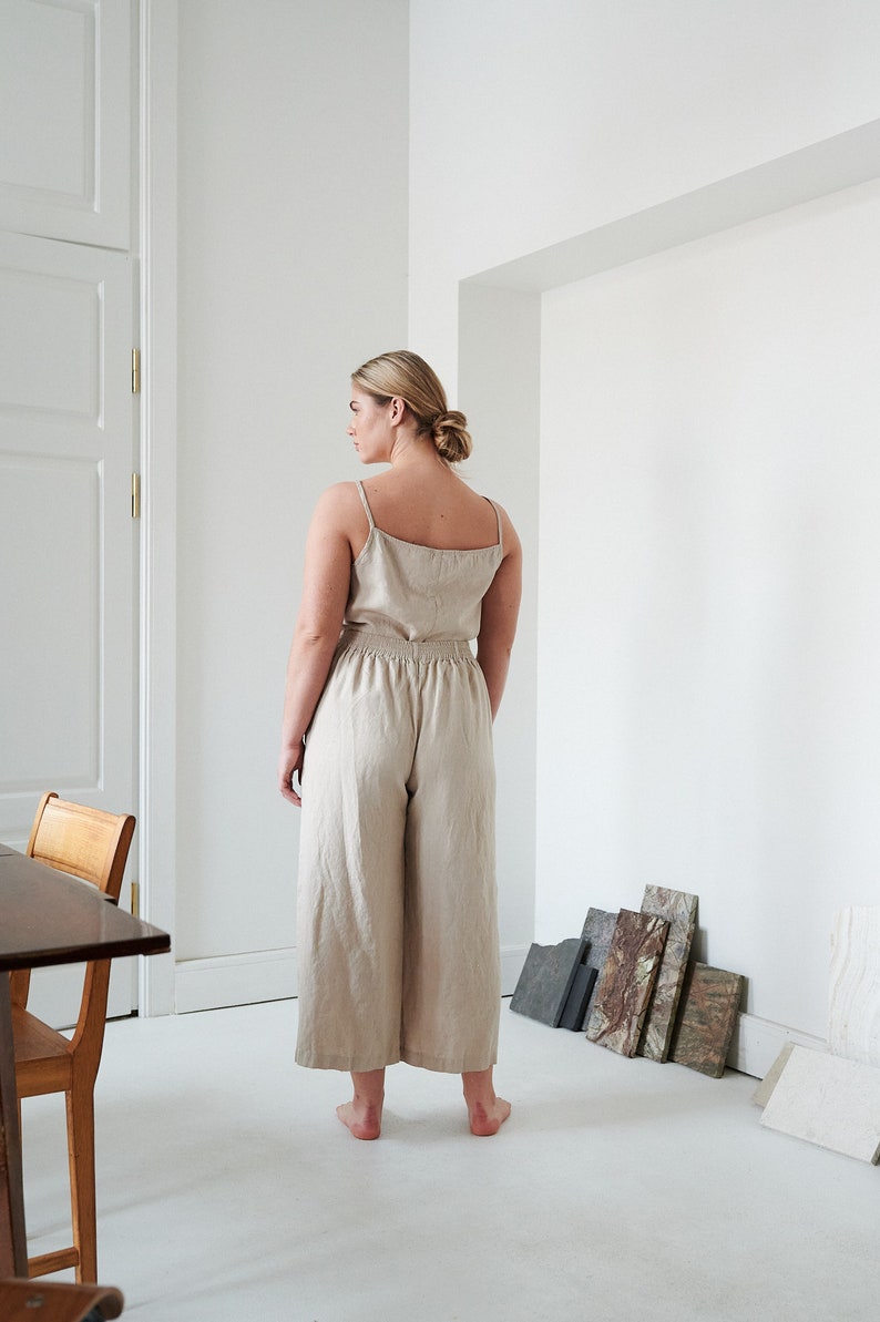 May include: A woman wearing a beige linen jumpsuit with a wide leg silhouette. The jumpsuit has a scoop neck and thin straps. The woman is standing in a white room with a wooden table and chairs in the background.
