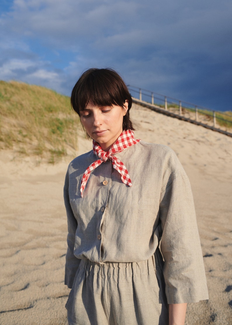 May include: A woman wearing a light gray linen jumpsuit with a red and white gingham scarf tied around her neck. She is standing on a sandy beach with a grassy dune in the background.