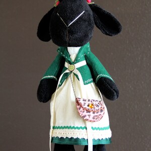 May include: A black plush sheep wearing a green jacket, a white dress with green and white trim, and a leopard print purse. The sheep has red and green flowers on its head.