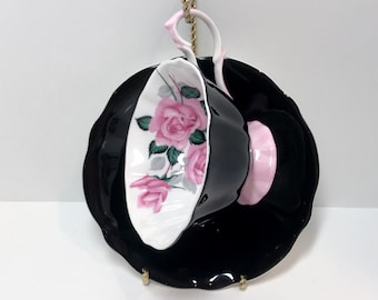 May include: A black teacup and saucer set with a pink rose floral design. The teacup has a white interior and the saucer has a black exterior with a pink rim.