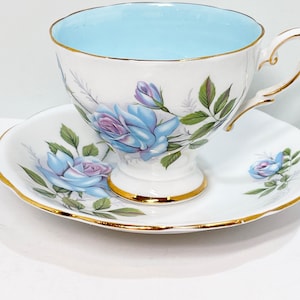 May include: A white teacup and saucer set with a blue and purple floral design. The cup has a light blue interior and a gold rim. The saucer has a scalloped edge and a gold rim.