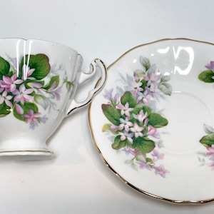 May include: A white teacup and saucer with a floral pattern. The pattern features pink and purple flowers with green leaves on a white background. The cup and saucer have a gold trim.