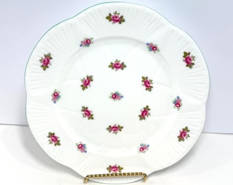 May include: A white ceramic plate with a scalloped edge and a thin green rim. The surface features a repeating pattern of small pink rose clusters with green leaves and tiny blue flowers arranged in a symmetrical grid across the center.