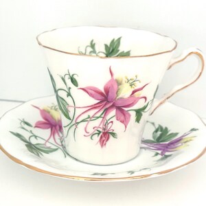 Puede incluir: Juego de taza y platillo blancos con diseño floral. La taza y el platillo están adornados con flores rosas y moradas, hojas verdes y ribetes dorados. La taza tiene un asa curvada y un borde festoneado.