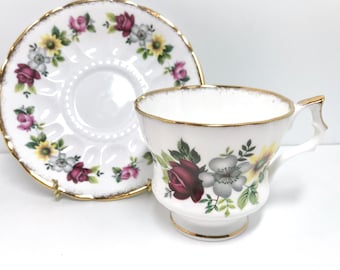 May include: A white teacup and saucer set with a floral pattern featuring red, yellow, and gray flowers. The set has a gold trim around the edges.