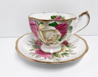 May include: A white teacup and saucer with a gold rim. The cup and saucer are decorated with a floral pattern of pink, white, and red roses.