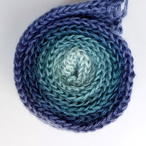 Pode incluir: Uma bola de fio em um degrad&ecirc; de cores azul, verde e branco. O fio est&aacute; enrolado em um c&iacute;rculo e est&aacute; pronto para ser usado para tricotar ou fazer croch&ecirc;.