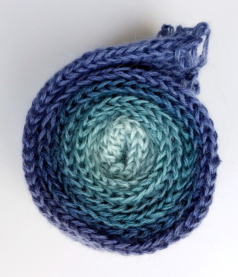 Pode incluir: Uma bola de fio em um degrad&ecirc; de cores azul, verde e branco. O fio est&aacute; enrolado em um c&iacute;rculo e est&aacute; pronto para ser usado para tricotar ou fazer croch&ecirc;.