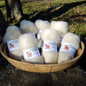 Puede incluir: Una cesta de mimbre llena de bolas de lana blancas y esponjosas. Cada bola tiene una etiqueta con el texto "Amity Creek Fiber P.O. Box 100 Duluth, MN www.amitycreekfiber.com".