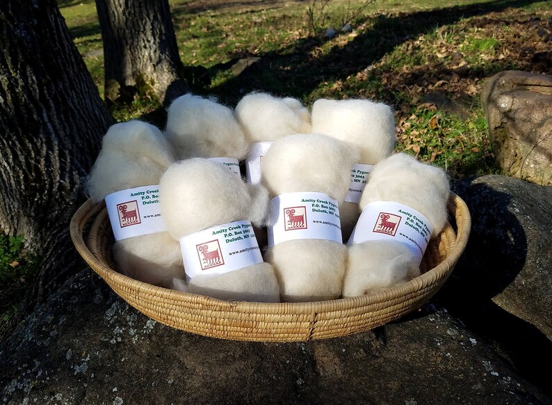 Puede incluir: Una cesta de mimbre llena de bolas de lana blancas y esponjosas. Cada bola tiene una etiqueta con el texto "Amity Creek Fiber P.O. Box 100 Duluth, MN www.amitycreekfiber.com".