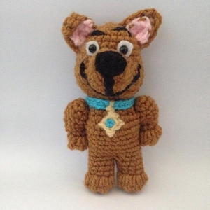 Puede incluir: Un juguete de peluche de Scooby Doo, un perro de dibujos animados, hecho a crochet de color marrón. El juguete tiene un collar azul con una estrella amarilla y una gema turquesa en el centro. El juguete tiene una gran sonrisa y ojos grandes.