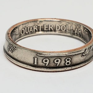 May include: A silver ring made from a quarter dollar coin. The ring features the text "QUARTER DOLLAR" and the year "1998" engraved on its surface. The ring has a worn, vintage appearance.