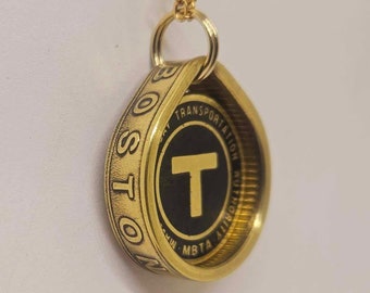 May include: Gold-colored pendant necklace featuring a Boston MBTA token. The token has a black center with a gold "T" symbol and the words "MBTA" and "TRANSPORTATION AUTHORITY". The pendant is suspended from a delicate gold chain.
