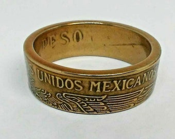 May include: A gold-toned ring with the words "PESO" and "ESTADOS UNIDOS MEXICANOS" engraved on the surface. The ring features an eagle emblem, showcasing a vintage or antique aesthetic. The ring is a jewelry item.