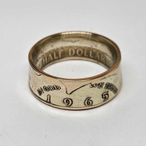 May include: A gold-toned ring made from a 1965 half dollar coin. The ring features the inscription "In God We Trust" and "Half Dollar" on the band.