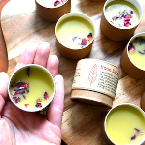 Può includere: Primo piano di diversi piccoli contenitori rotondi riempiti con un balsamo giallo pallido e adornati con petali di rosa secchi. Un contenitore è tenuto in mano. È visibile anche un piccolo contenitore cilindrico con il testo "Moon Balm".