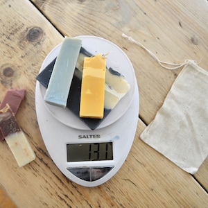 Puede incluir: Una báscula de cocina digital blanca con una pantalla negra que muestra 131 gramos. Hay cuatro barras de jabón en la báscula, una azul, una negra, una amarilla y una marrón y blanca.