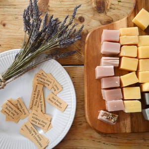 May include: A wooden board presents soap samples in yellow, pink, grey, and black. A white plate holds small tags with soap names. A bundle of dried lavender sits beside the plate. The tags read "Patchouli & Cypress", "Black Rose", and "Sea Song".