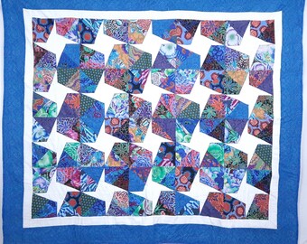 May include: A colorful quilt with a geometric pattern. The quilt features a white center with blue, purple, and floral patterned shapes. It has a white border and a blue patterned edge.