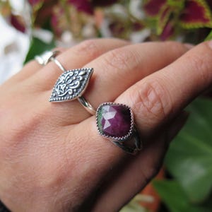 May include: Two silver rings are displayed on a hand. One ring features a detailed, leaf-shaped design. The other ring has a square, faceted, deep red gemstone. Both rings have a detailed silver band.