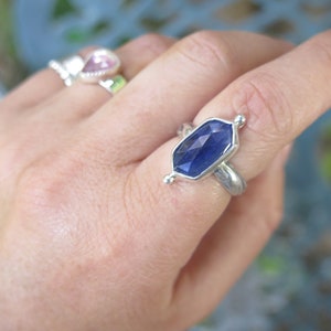 Op de afbeelding: Een zilveren ring met een diepblauwe edelsteen gezet in een zeshoekige vorm. De ring heeft een getextureerde band en wordt gedragen aan een vinger.
