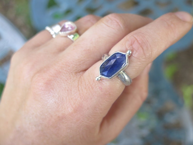 Op de afbeelding: Een zilveren ring met een diepblauwe edelsteen gezet in een zeshoekige vorm. De ring heeft een getextureerde band en wordt gedragen aan een vinger.