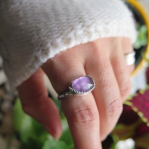 May include: A silver ring with a purple gemstone. The ring is on a person's finger.