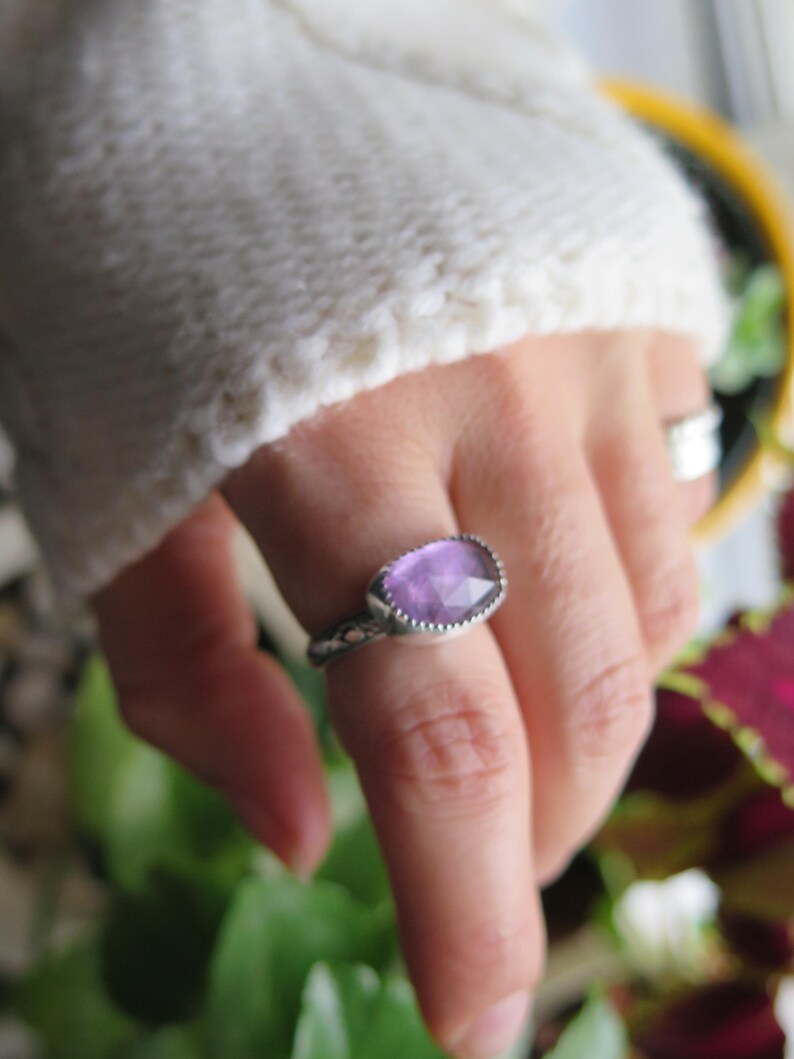 May include: A silver ring with a purple gemstone. The ring is on a person's finger.