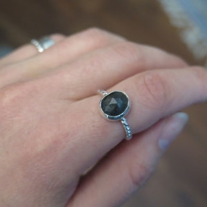 K&ouml;nnte beinhalten: Ein silberner Ring mit einem dunkelgrauen Edelstein. Der Ring hat ein gedrehtes Band.
