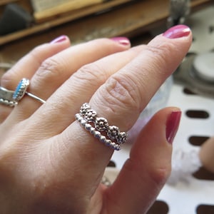 May include: Two silver rings on a finger. The top ring has a design of small circles with a raised centre. The bottom ring has a design of small beads.