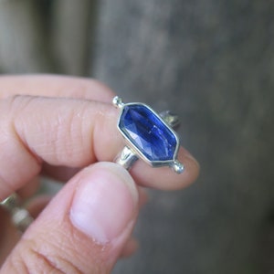 Op de afbeelding: Een zilveren ring met een blauwe edelsteen in een zeshoekige vorm. De ring heeft kleine zilveren accenten aan de zijkanten.