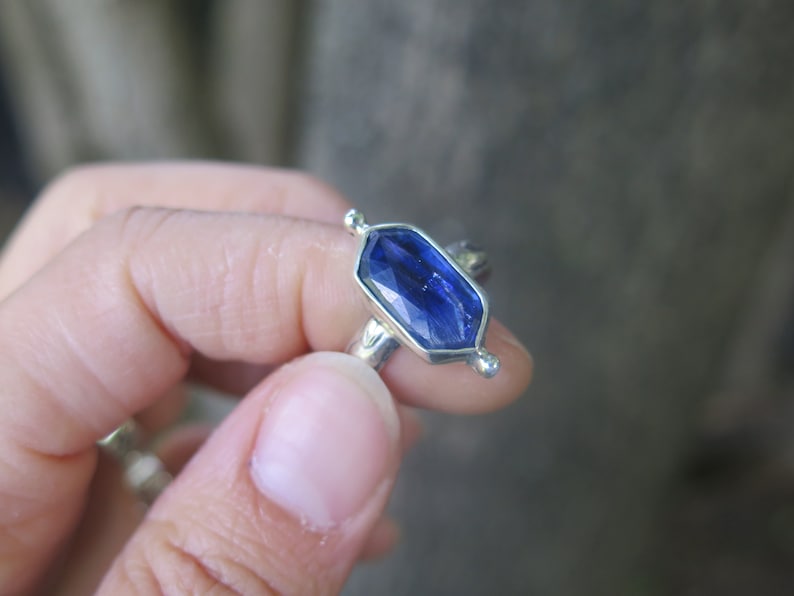 Op de afbeelding: Een zilveren ring met een blauwe edelsteen in een zeshoekige vorm. De ring heeft kleine zilveren accenten aan de zijkanten.