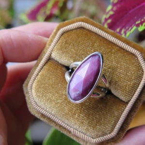 May include: A silver ring featuring a faceted, marquise-shaped deep magenta gemstone set in a smooth metal bezel. The ring is displayed in a square, mustard-yellow velvet ring box with a central slot for the band, showcasing the stone's texture.