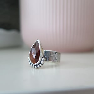 May include: A silver ring with a teardrop-shaped, faceted, reddish-brown gemstone. The ring has a sun and moon design on the band and a beaded detail at the base of the gemstone. The ring is on a white surface.