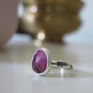 May include: A silver ring featuring a large, oval-cut, magenta gemstone. The ring's band has a decorative leaf pattern. The ring is set against a blurred background with a pink vase and a gold-coloured object.