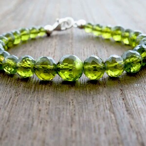 May include: A bracelet with faceted, olive-green gemstone beads. The beads are strung together with a silver clasp. The bracelet is displayed on a wooden surface, highlighting the vibrant colour and sparkle of the gemstones.
