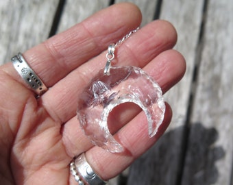 May include: A clear quartz crescent moon pendant on a silver chain. The pendant is held in a hand.