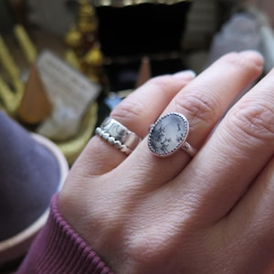 May include: A silver ring with an oval, dendritic agate stone. The stone has a grey and black pattern. A second silver ring with a beaded design is also visible. The rings are on a hand.