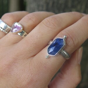 Op de afbeelding: Een zilveren ring met een blauwe edelsteen in een zeshoekige vorm. De ring zit aan een vinger.