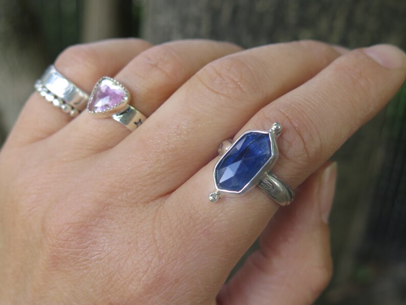 Op de afbeelding: Een zilveren ring met een blauwe edelsteen in een zeshoekige vorm. De ring zit aan een vinger.