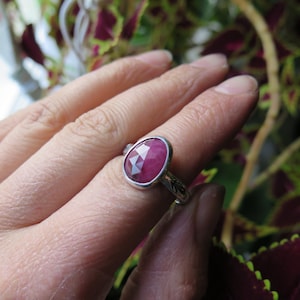 May include: A silver ring with a large, faceted, oval-shaped ruby gemstone. The ruby is a deep pinkish-red color. The ring has a detailed leaf design on the band.