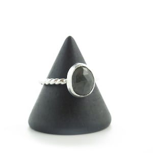 K&ouml;nnte beinhalten: Ein silberner Ring mit einem gedrehten Band und einem dunkelgrauen Edelstein. Der Ring wird auf einem schwarzen kegelf&ouml;rmigen St&auml;nder pr&auml;sentiert.